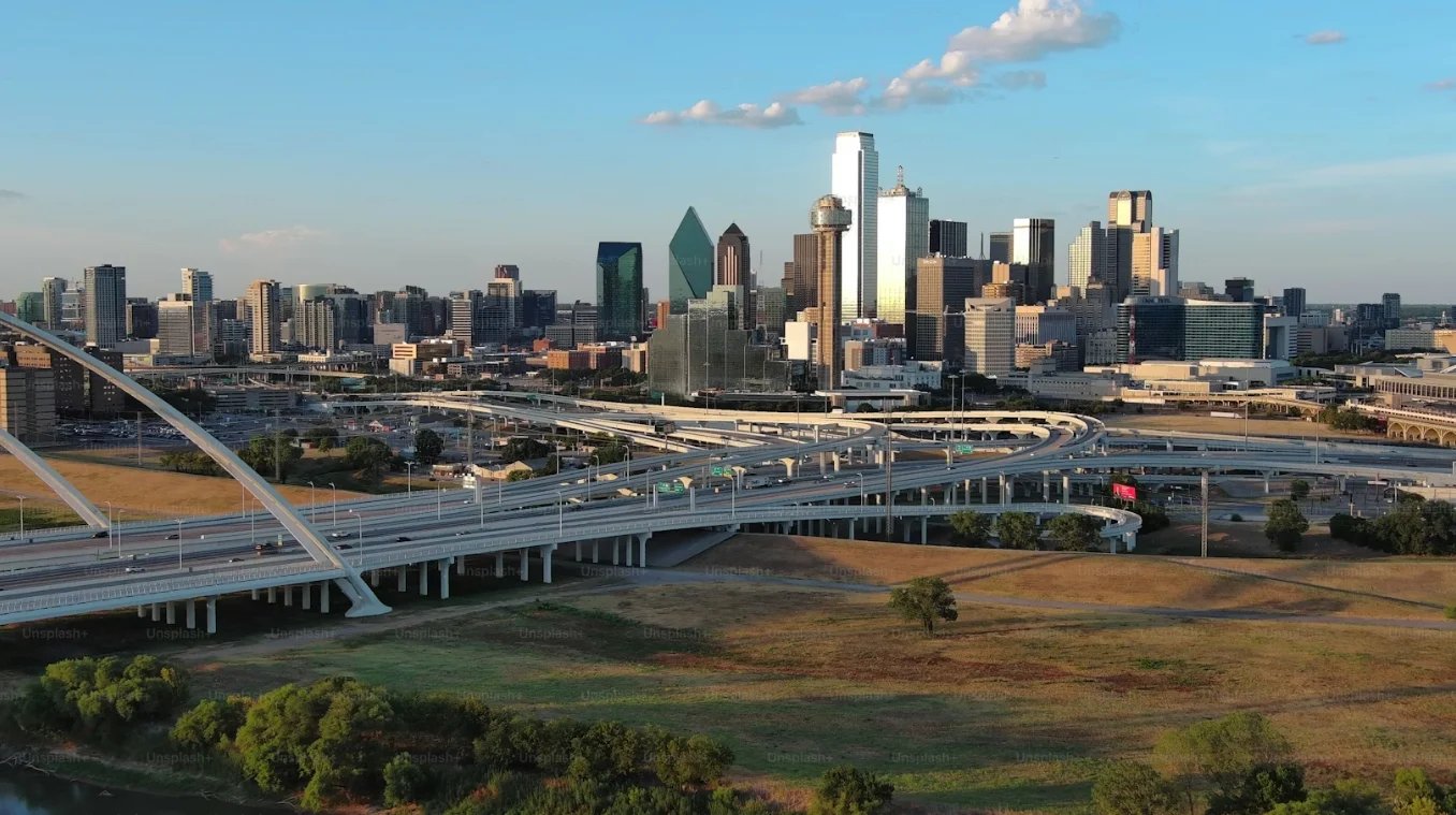 Dallas skyline with downtown buildings and highway interchange spanning grassy terrain below under blue sky