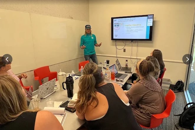 Instructor in green shirt presenting to group of women seated at table with laptops and water bottles, with presentation screen on wall behind