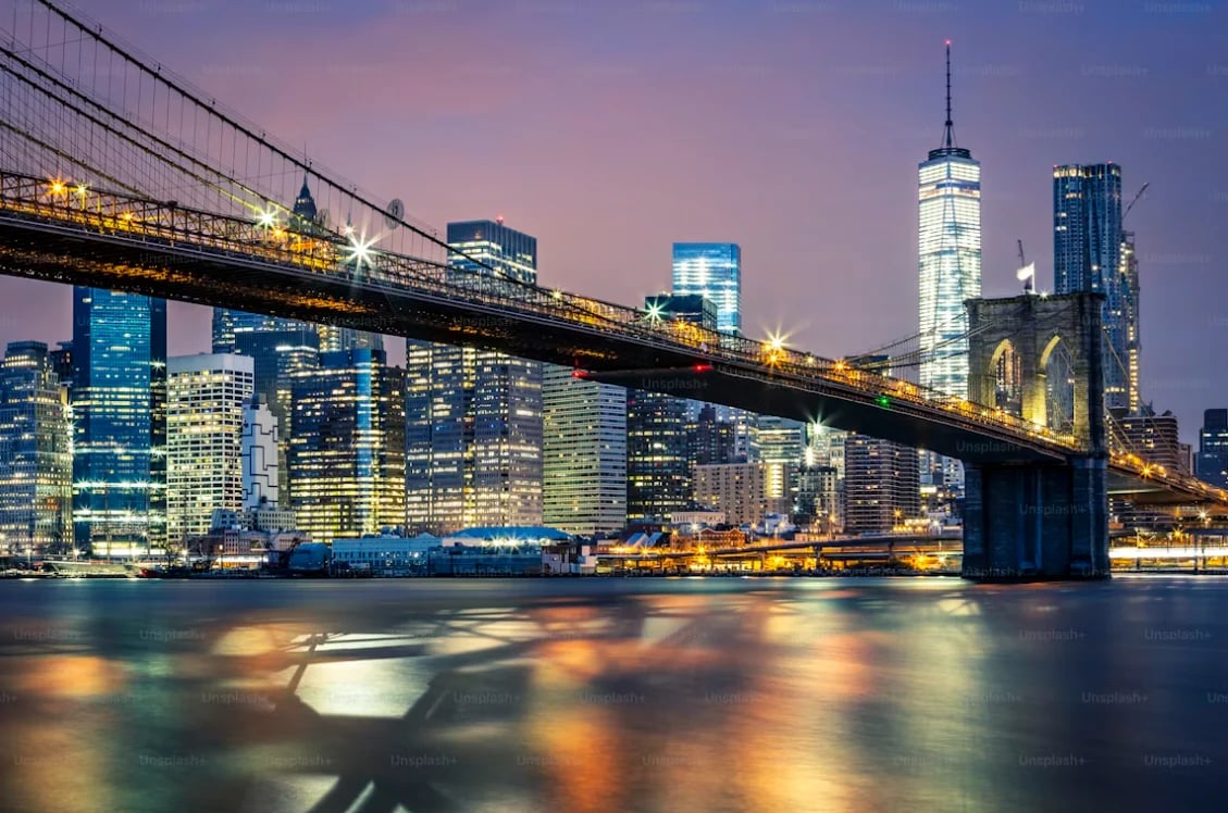 Brooklyn Bridge spanning the East River at twilight with illuminated Manhattan skyline and skyscrapers reflected in the water