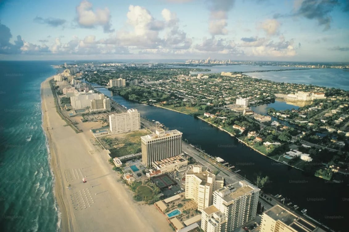 Aerial view of a coastal city with beachfront high-rise buildings, waterways, and residential neighborhoods under a blue sky with white clouds