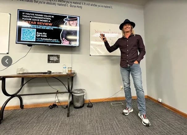 Person in dark shirt and jeans presenting in a classroom with a monitor on the wall showing a QR code and presentation materials