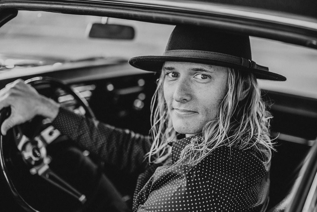 Black and white portrait of Bobby Kerr with long blonde hair wearing a black cowboy hat and polka dot jacket, sitting in a car