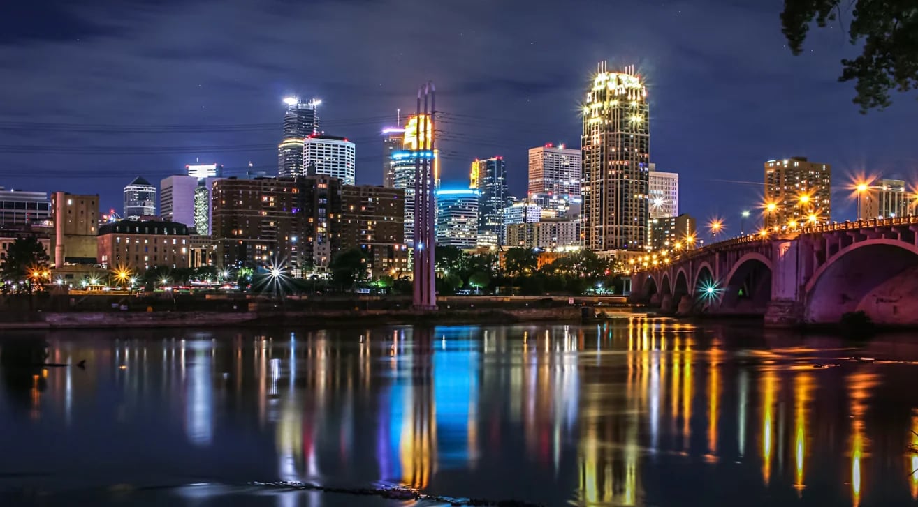 Minneapolis skyline at dusk with illuminated buildings reflected in the Mississippi River and Stone Arch Bridge
