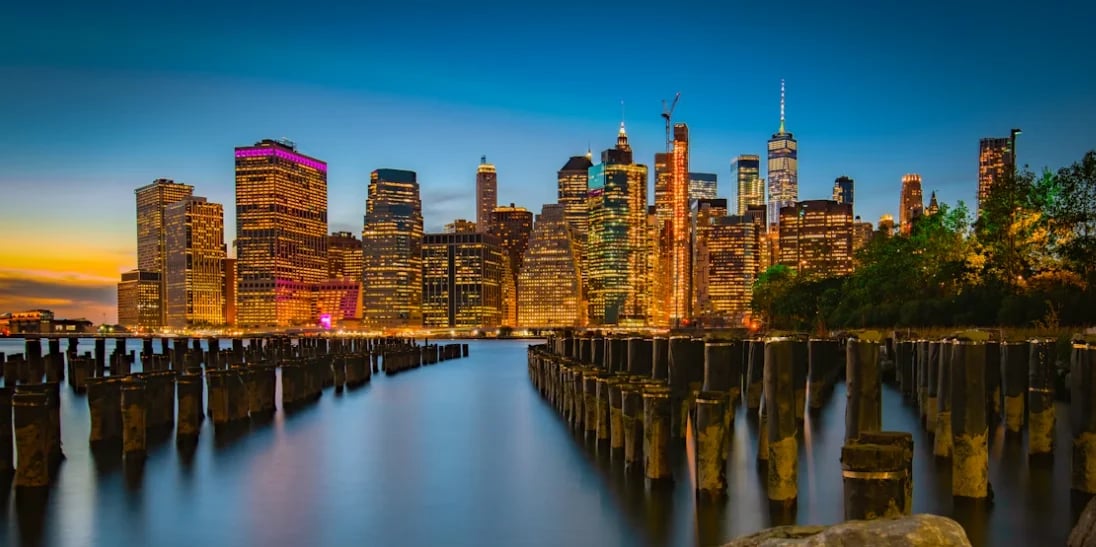 Manhattan skyline illuminated at twilight with wooden pilings reflecting in calm water