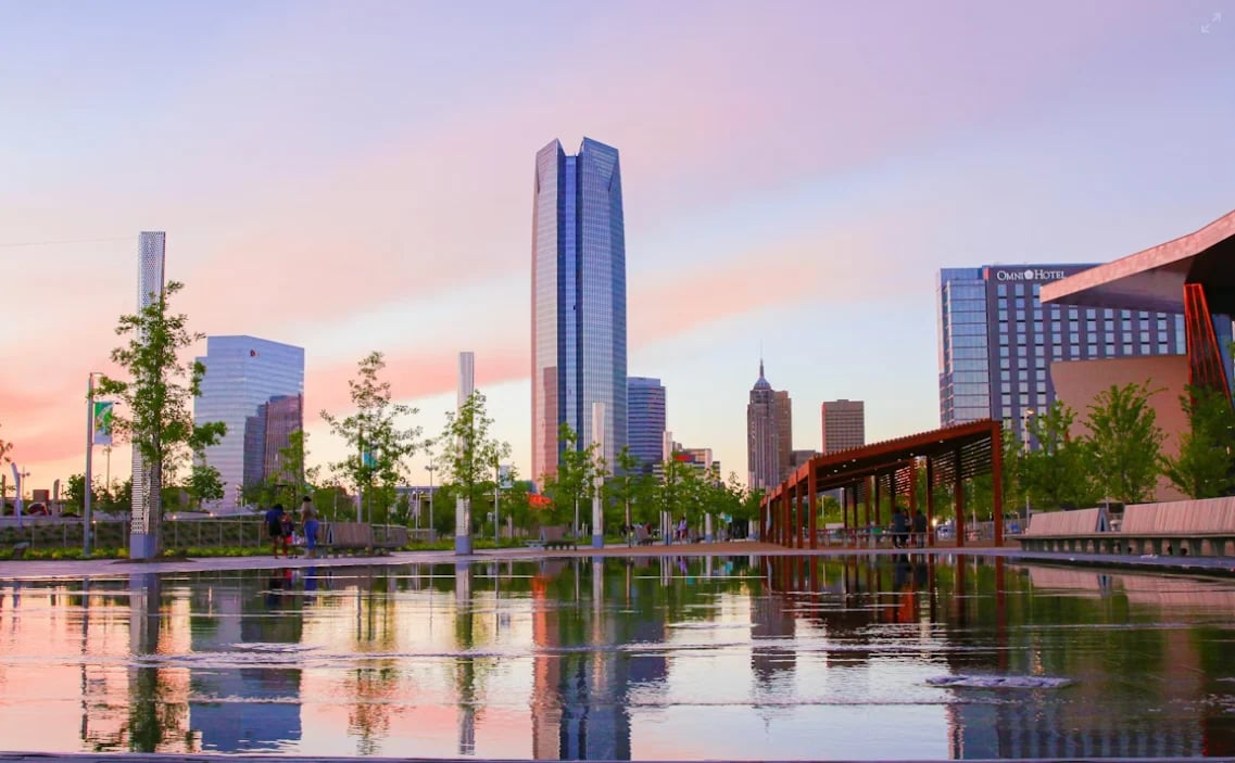 Urban skyline with tall buildings reflected in a calm water channel at sunset, lined with green trees and modern architecture
