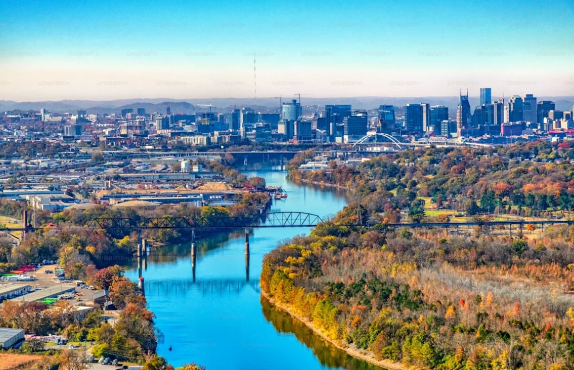 Aerial view of a city skyline with a turquoise river, autumn trees, and a bridge in the foreground