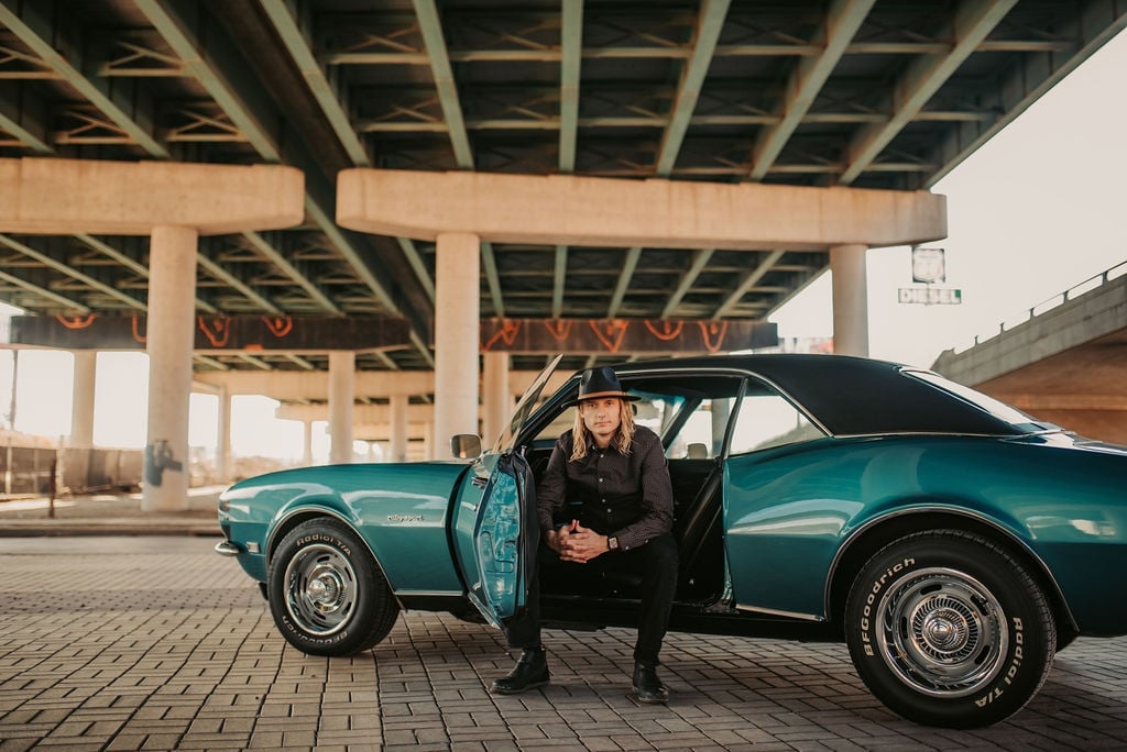 Man in black suit posing with vintage turquoise car under bridge overpass