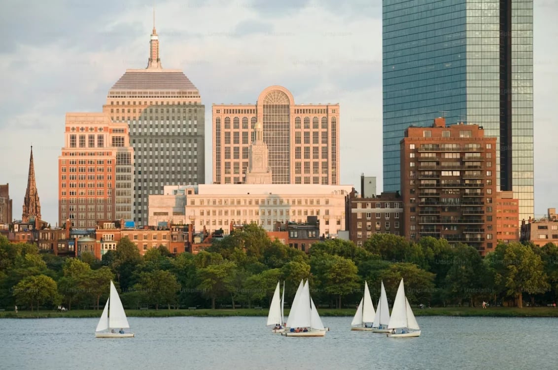 Three white sailboats on water with Boston skyline and historic brick buildings in the background